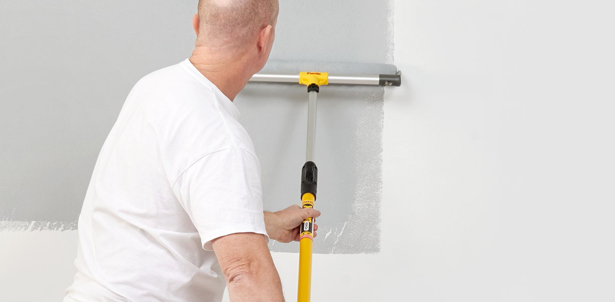 Pro painter using a large roller frame, a roller and an extension pole to apply gray paint to an interior wall.
