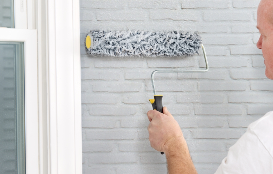Pintor profesional aplicando pintura con rodillo grande en pared de ladrillo exterior.
