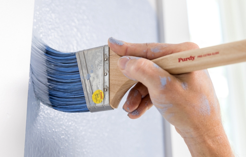 Painter cutting in around the window trim with light blue paint using a synthetic paint brush.