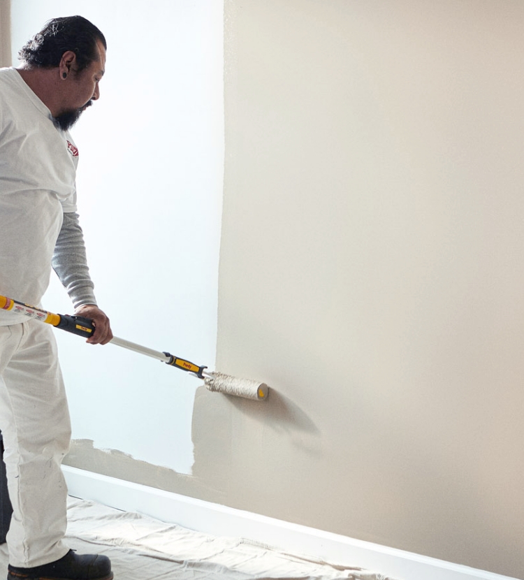 Pro painter covering a large wall with neutral paint color using a roller and an extension pole.