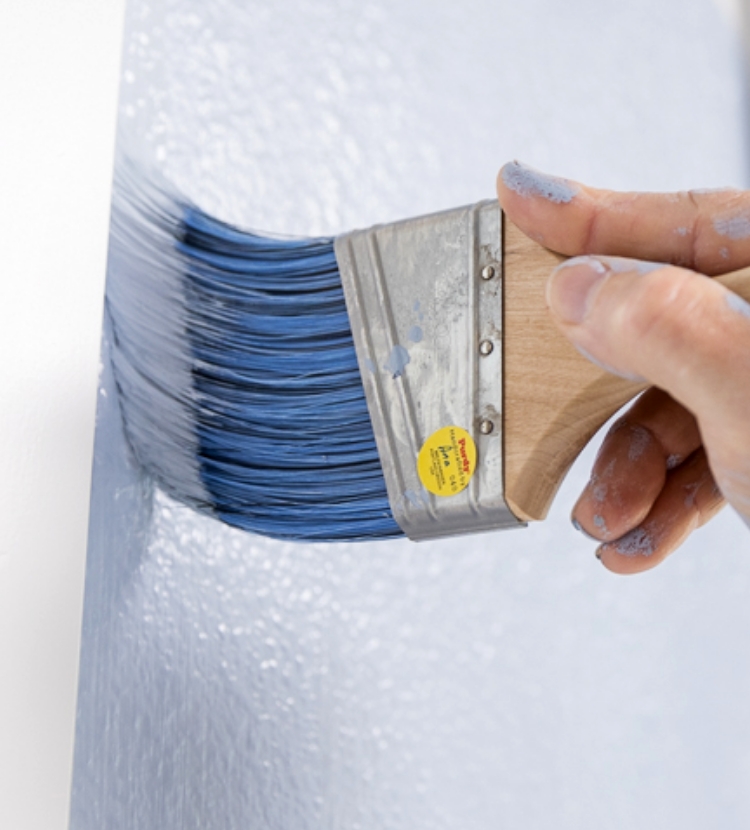 Painter cutting in around the window trim with light blue paint using a synthetic paint brush.