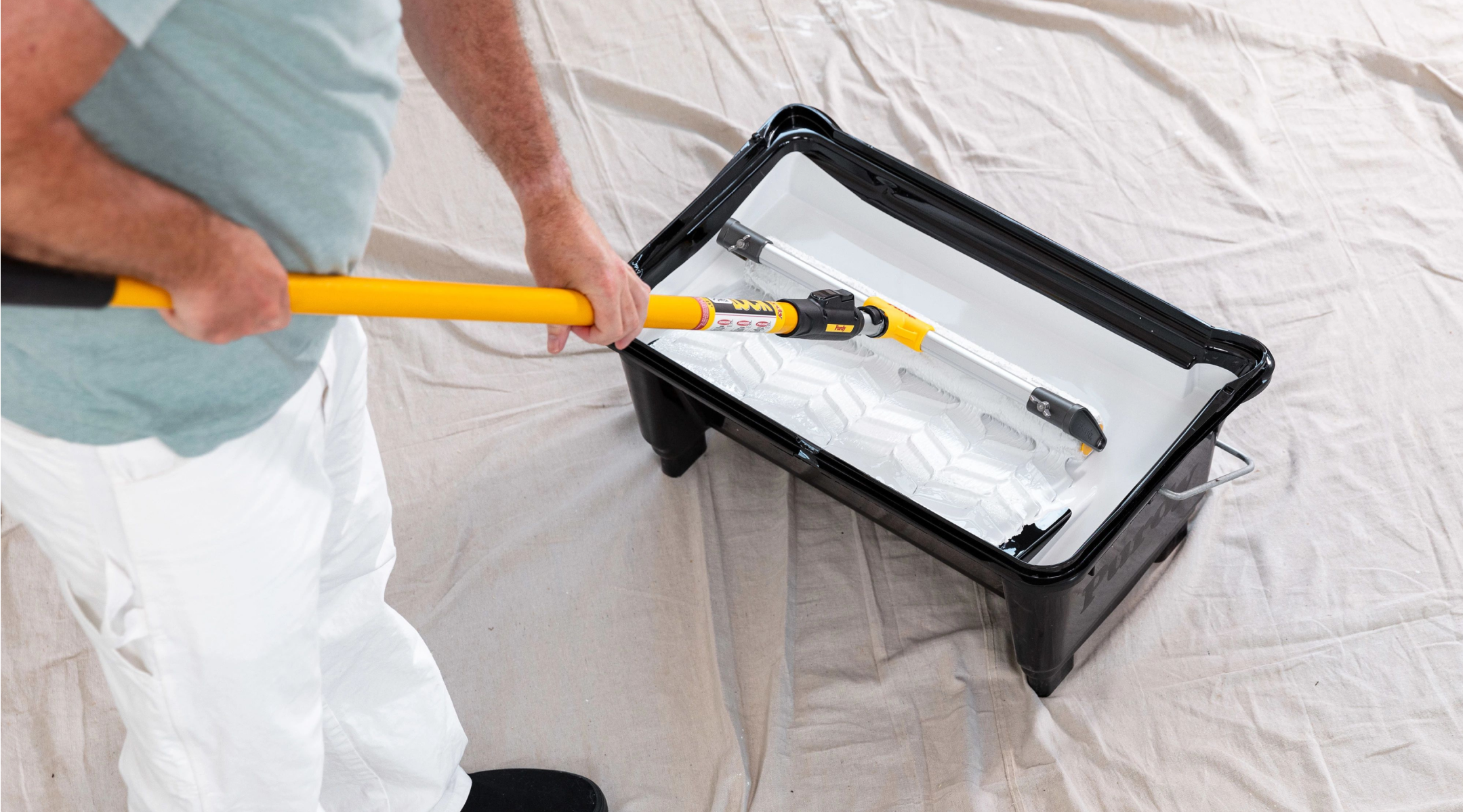 Painter rolling out white paint with a large roller in an eighteen-inch paint tray.